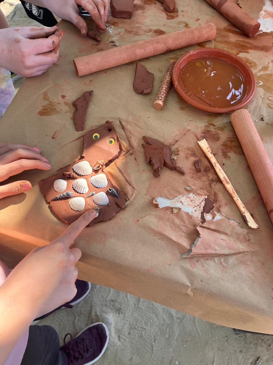 Manos de varios niños modelando arcilla y decorándola con conchas sobre la mesa del taller