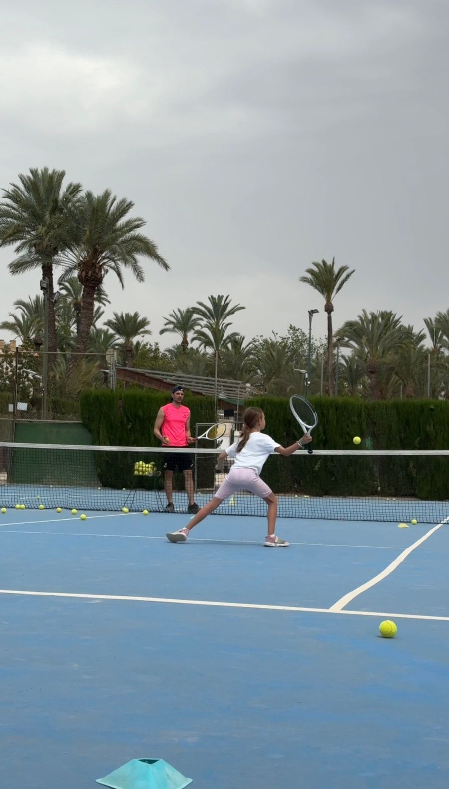Niña golpeando la pelota con la raqueta en la pista de tenis, con el monitor al otro lado de la red y las palmeras al fondo