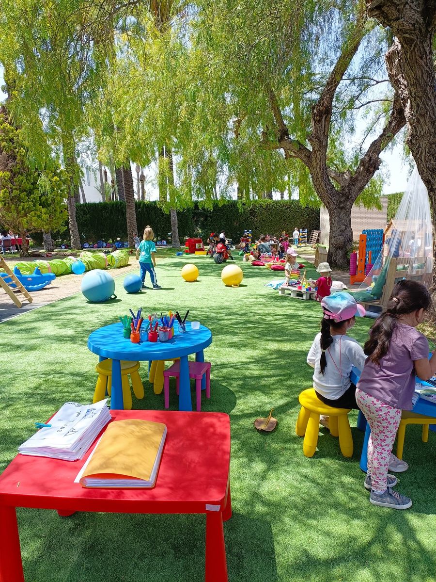 Jardín de césped con mesitas y taburetes de colores, pelotas gigantes y juguetes, donde varios niños de Infantil dibujan y juegan a la sombra